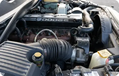A Dodge Ram engine compartment is shown with the hood open. The engine is visible with various components such as hoses, belts, and the air intake system. The Dodge logo is prominent on the engine cover.