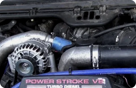 A close-up view of a truck engine compartment, focusing on the turbo diesel components. A label reading "POWER STROKE V8 TURBO DIESEL" is visible in the foreground. Various hoses, belts, and mechanical parts are tightly arranged within the engine bay. The lighting highlights the textures and metallic surfaces of the engine components.