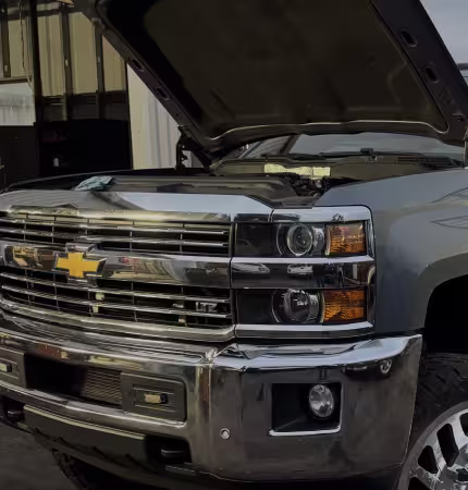 A silver 2015 Chevrolet Silverado 2500HD pickup truck is shown with its hood open, parked indoors. The truck's front grille, headlights, and bumper are clearly visible. The open hood reveals a portion of the engine compartment. The setting appears to be a garage or workshop.