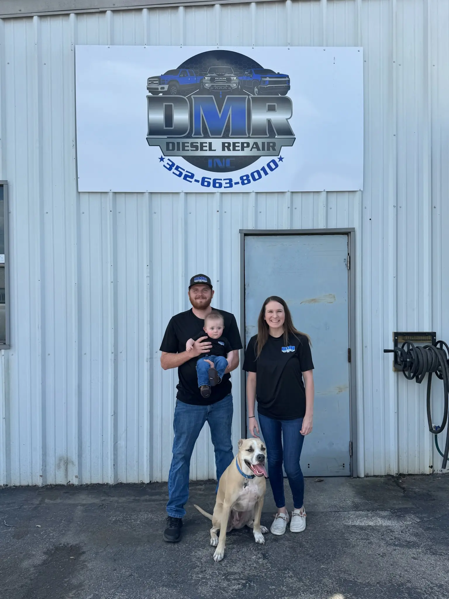 A family of three, including a baby, stands with their dog in front of DMR Diesel Repair.