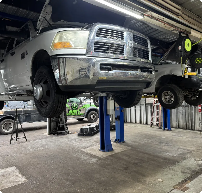 A light-colored Ram truck is elevated on a blue hydraulic lift in an auto repair shop. The truck's hood is open, and the front is facing the viewer. Another vehicle is visible on a lift in the background, along with various tools and equipment typical of an automotive service environment.