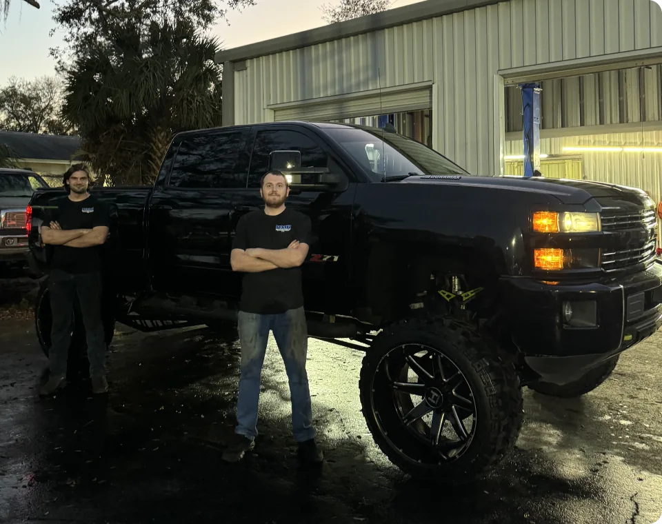 Two mechanics standing in front of a dark-colored pickup truck. Both are wearing dark shirts with a logo on them. The truck is partially visible, with its headlights and grille prominent.