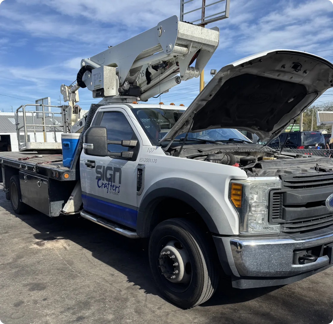 A white 2019 Ford F-550 XLT Super Duty truck, equipped with a Versalift bucket lift, is parked outdoors with its hood open. The truck features a flatbed, utility boxes, and the "SIGN Crafters" logo on the driver's side door. The bucket lift arm is extended upwards. The sky is partly cloudy.