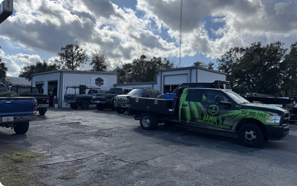 A collection of pickup trucks are parked in a lot outside several light-colored, metal buildings, possibly a business or service center. The truck in the foreground is a black flatbed truck with green graphics that read "Jamie's Landscaping & Pavers" along with a phone number. The sky is overcast with a mix of dark and light clouds. Trees are visible in the background, suggesting a suburban or rural setting.