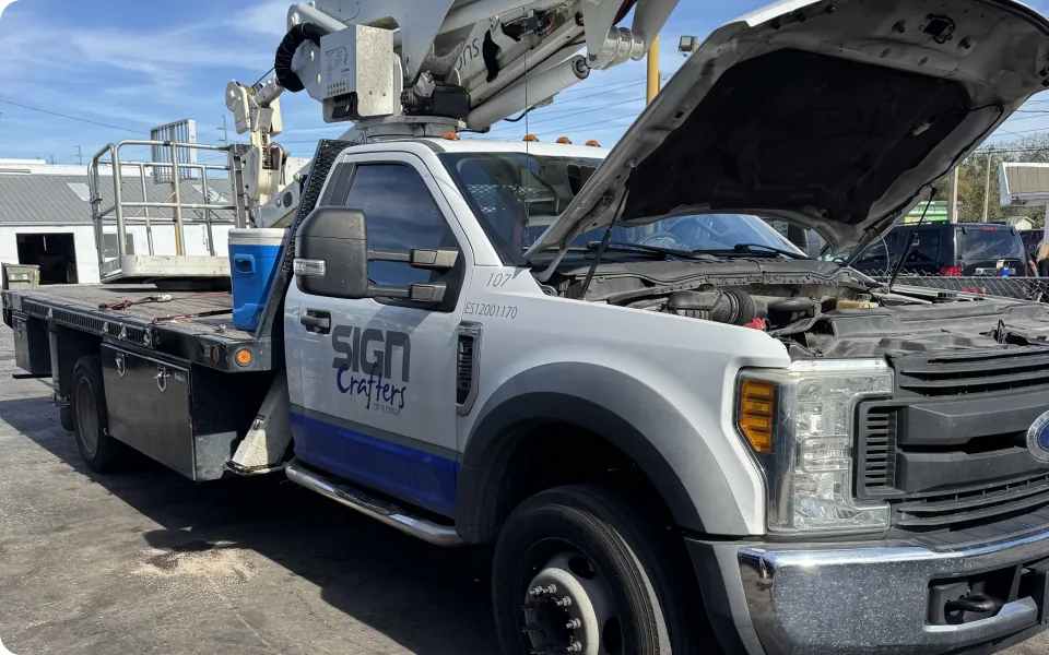 A white 2019 Ford F-550 XLT Super Duty truck, equipped with a Versalift bucket lift, is parked outdoors with its hood open. The truck features a flatbed, utility boxes, and the "SIGN Crafters" logo on the driver's side door. The bucket lift arm is extended upwards. The sky is partly cloudy.