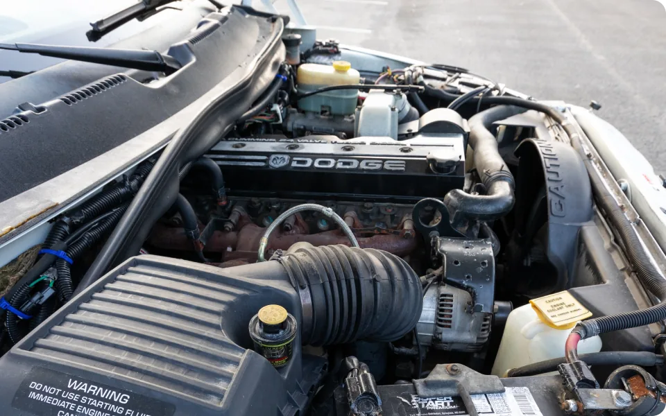 A Dodge Ram engine compartment is open, revealing the engine with its various components, including hoses, belts, and the air intake system. The "DODGE" logo is visible on the engine cover.