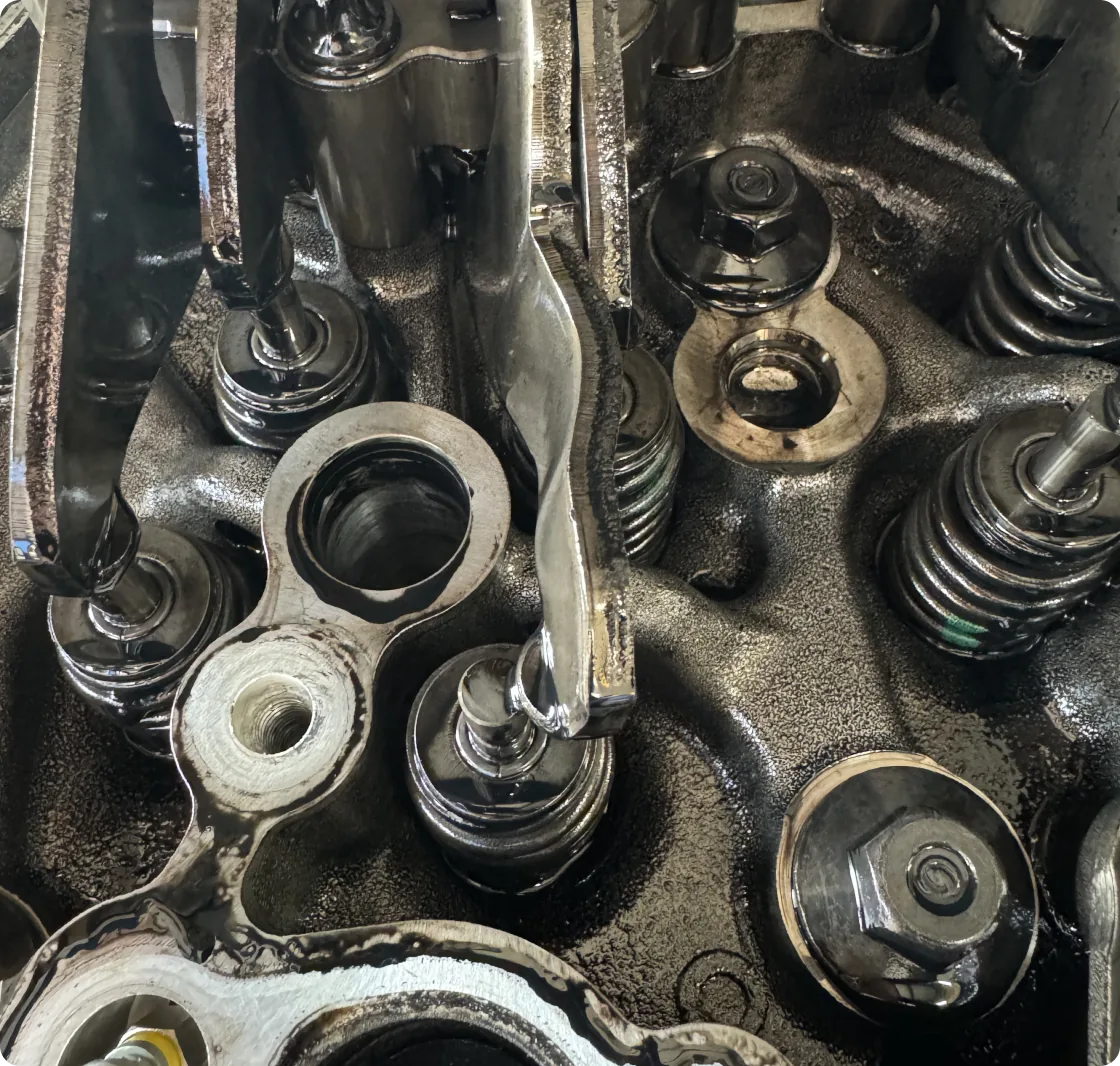 A detailed close-up of an automotive engine's internal components, likely within the cylinder head, showcasing valves, springs, and rocker arms. The parts are metallic and appear lubricated.