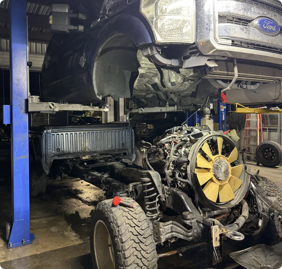 A Ford truck chassis with the body detached is lifted on a blue hydraulic lift in a repair shop. The engine with a yellow fan, wheels, and frame are clearly visible.