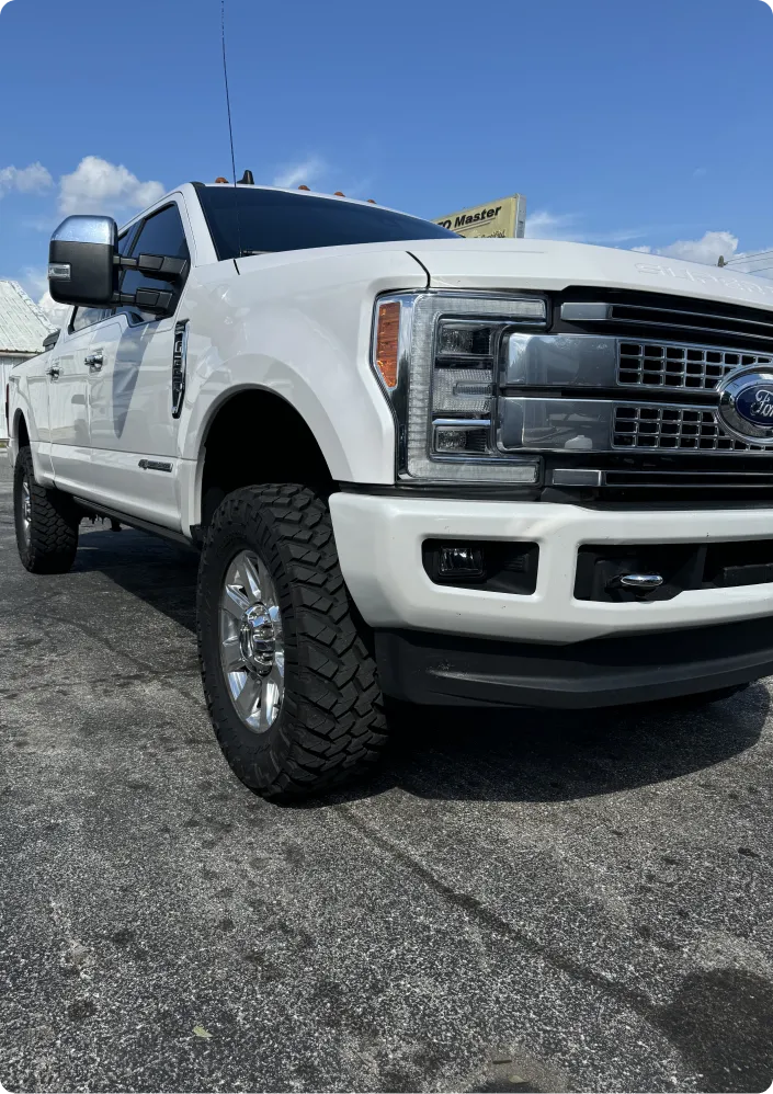 A white Ford F-250 Platinum pickup truck parked on an asphalt surface under a clear blue sky with scattered clouds. The truck is viewed from a low angle, emphasizing its front grille, headlights, and chrome wheels. The "Platinum" badging is visible on the side.